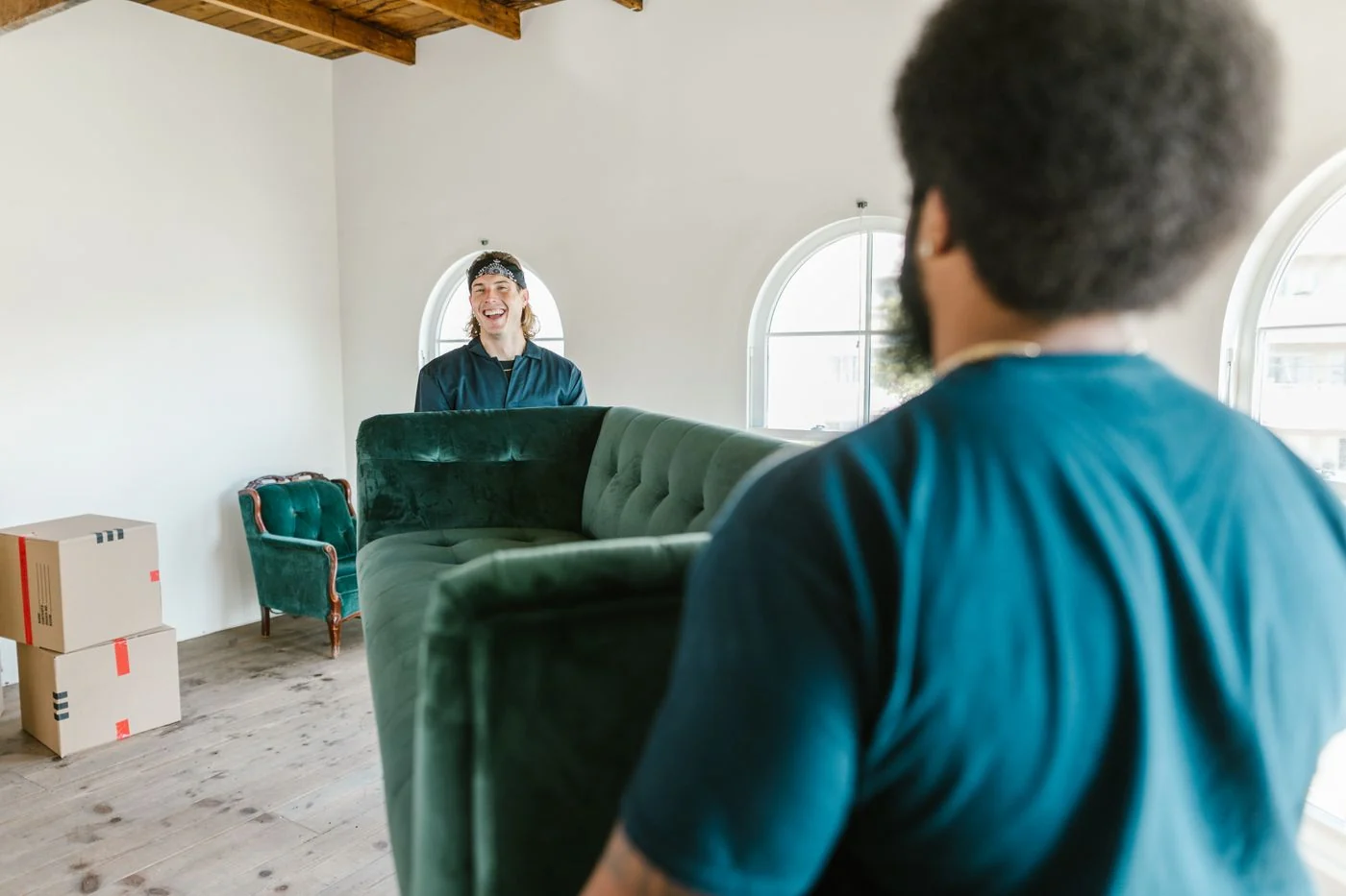 Two movers sharing the work of carrying a green sofa through a sunlit home.